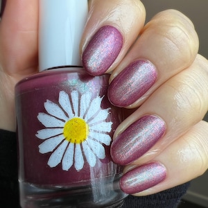 May include: A clear glass bottle of nail polish with a white daisy on the label. The polish is a deep burgundy color with a shimmer finish. The bottle is being held by a hand with nails painted in the same color polish.