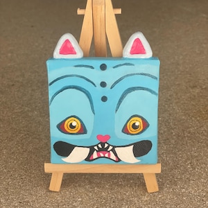 May include: A small, square painting of a cartoon cat face with a light blue background. The cat has large yellow eyes, white fangs, and pink accents. The painting is displayed on a small wooden easel.