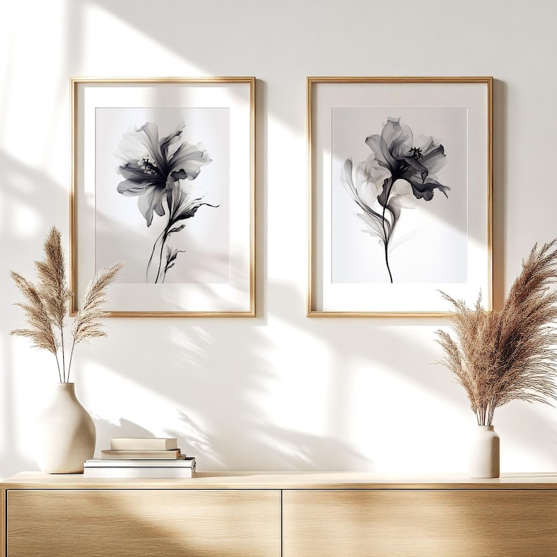 May include: Two framed floral art prints. Each print features a black and white flower illustration with delicate details. The frames are gold-colored, and the prints are displayed on a white wall above a wooden cabinet. Decorative vases with dried plants are on the cabinet.