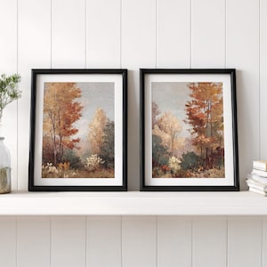 May include: Two framed landscape paintings. Each features a forest scene with trees in shades of red, orange, and yellow, suggesting autumn. The paintings are in black frames and rest on a white shelf.