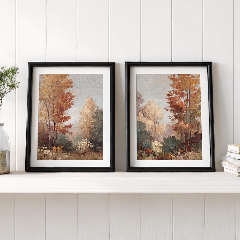 May include: Two framed landscape paintings. Each features a forest scene with trees in shades of red, orange, and yellow, suggesting autumn. The paintings are in black frames and rest on a white shelf.