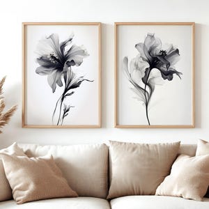 May include: Two framed floral art prints. Each print features a grayscale image of a large flower with delicate, translucent petals and dark centers. The prints are in light wood frames, displayed above a beige sofa with matching pillows.