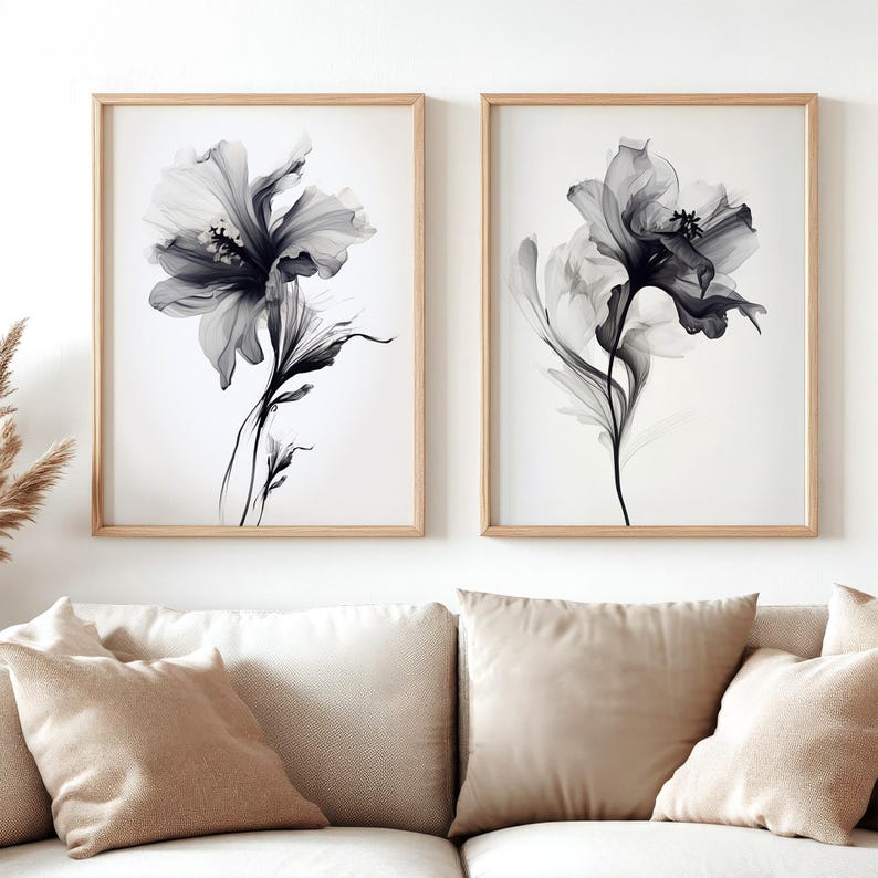 May include: Two framed floral art prints. Each print features a grayscale image of a large flower with delicate, translucent petals and dark centers. The prints are in light wood frames, displayed above a beige sofa with matching pillows.