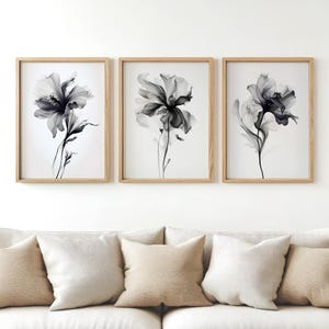 May include: Three framed floral art prints. Each print features a grayscale image of a flower with delicate petals and stems against a white background. The frames are made of light-colored wood. The artwork is displayed above a sofa with neutral-colored pillows.