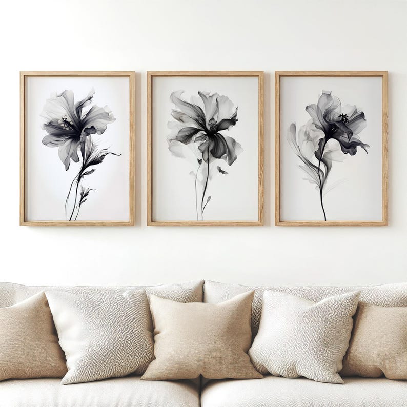 May include: Three framed floral art prints. Each print features a grayscale image of a flower with delicate petals and stems against a white background. The frames are made of light-colored wood. The artwork is displayed above a sofa with neutral-colored pillows.