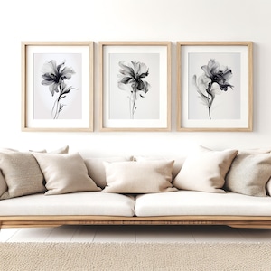 May include: Three framed floral art prints. Each print features a grayscale watercolor of a flower with delicate petals and stems. The frames are light wood, and the prints are displayed above a beige sofa with matching pillows.