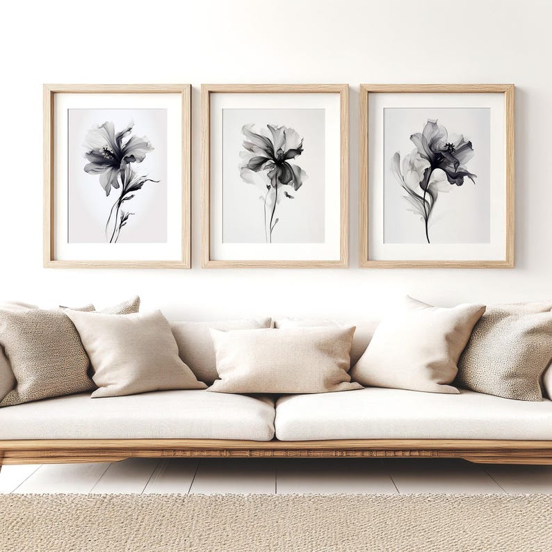 May include: Three framed floral art prints. Each print features a grayscale watercolor of a flower with delicate petals and stems. The frames are light wood, and the prints are displayed above a beige sofa with matching pillows.
