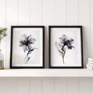 May include: Two framed floral art prints. Each print features a grayscale image of a flower with delicate petals and stems. The frames are black and the background is white. The prints are displayed on a white shelf.