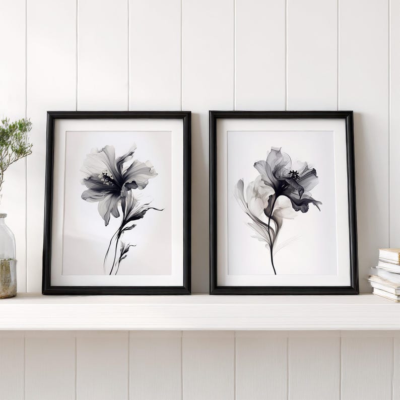 May include: Two framed floral art prints. Each print features a grayscale image of a flower with delicate petals and stems. The frames are black and the background is white. The prints are displayed on a white shelf.
