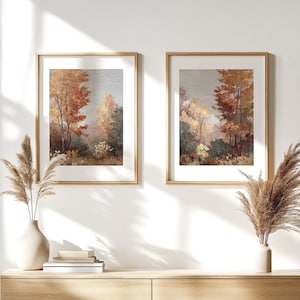 May include: Two framed landscape paintings with gold frames. The artwork depicts a forest scene with trees in shades of red, orange, and brown, suggesting an autumn theme. The paintings are displayed on a light-colored wooden surface.