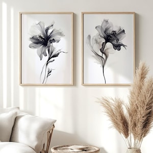 May include: Two framed floral art prints. Each print features a black and white, ethereal flower design against a white background. The frames are light wood. The artwork is displayed in a living room setting with neutral-toned furniture and decor.
