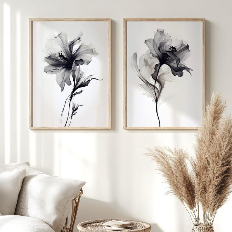May include: Two framed floral art prints. Each print features a black and white, ethereal flower design against a white background. The frames are light wood. The artwork is displayed in a living room setting with neutral-toned furniture and decor.