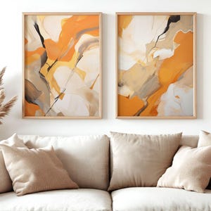 Puede incluir: Dos impresiones artísticas abstractas enmarcadas. Cada impresión presenta una mezcla de pinceladas naranjas, blancas, grises y negras. La obra de arte se exhibe sobre un sofá beige con cojines a juego, creando un diseño interior moderno y cohesivo.