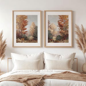 May include: Two framed landscape art prints. Each print features a forest scene with trees in shades of red, orange, and green. The artwork is displayed above a bed with white bedding and decorative pillows.
