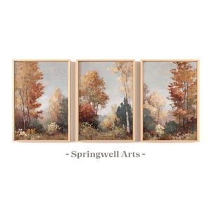 May include: Set of three framed landscape paintings. Each painting depicts a forest scene with trees in shades of orange, brown, and yellow, suggesting autumn. The frames are a light wood color, and the background is a soft, muted blue.
