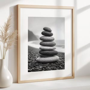 Puede incluir: Impresión enmarcada en blanco y negro de una pila de piedras lisas y redondeadas. Las piedras están apiladas unas sobre otras, creando una torre. El fondo muestra una playa y el océano.