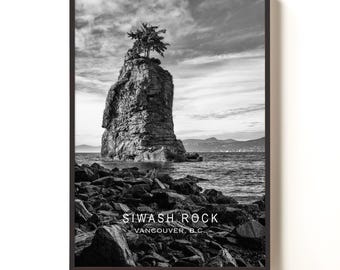May include: Monochrome framed print of Siwash Rock, Vancouver, B.C. The image depicts a tall rock formation with a tree on its summit, set against a cloudy sky and the sea. The foreground features a rocky beach.