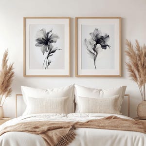 May include: Two framed floral art prints. Each print features a grayscale depiction of a flower with delicate petals and stems. The frames are light wood, and the prints are displayed above a bed with white bedding and decorative pillows.