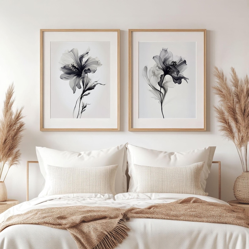 May include: Two framed floral art prints. Each print features a grayscale depiction of a flower with delicate petals and stems. The frames are light wood, and the prints are displayed above a bed with white bedding and decorative pillows.