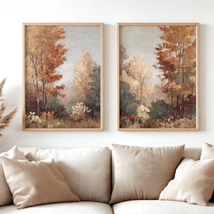 May include: Two framed landscape paintings depict an autumnal forest scene. The artwork features trees with red, orange, and yellow leaves, set against a soft blue sky. The paintings are in light wood frames, displayed above a beige sofa.