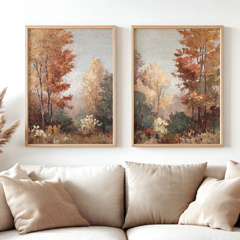 May include: Two framed landscape paintings depict an autumnal forest scene. The artwork features trees with red, orange, and yellow leaves, set against a soft blue sky. The paintings are in light wood frames, displayed above a beige sofa.