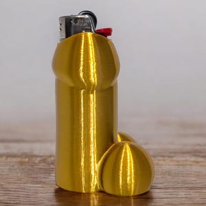 May include: A gold-colored lighter case with a phallic design. The case is made of a ribbed material and has a rounded base. A standard lighter is visible at the top, with a red button. The item is placed on a wooden surface.