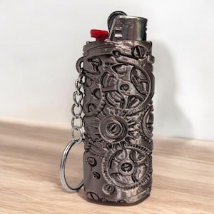 Puede incluir: Un encendedor gris oscuro con un dise&ntilde;o de engranajes de estilo steampunk. El encendedor tiene un bot&oacute;n de encendido rojo y un llavero plateado. El dise&ntilde;o presenta detalles intrincados de engranajes y elementos mec&aacute;nicos.