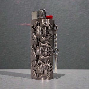 May include: A metallic gray lighter with a floral design. The lighter has a red ignition button and a small chain with a ring attached. The floral pattern is carved into the metal, adding texture and detail to the lighter's surface.