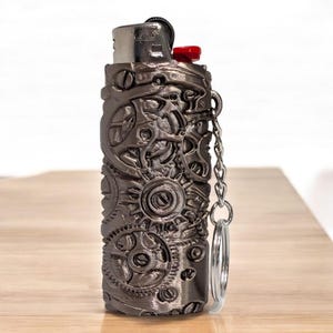 Puede incluir: Un encendedor gris met&aacute;lico con un bot&oacute;n de encendido rojo, encerrado en una cubierta decorativa con un dise&ntilde;o detallado de engranajes steampunk. Se adjunta una peque&ntilde;a cadena y un llavero. El encendedor est&aacute; sobre una superficie de madera.