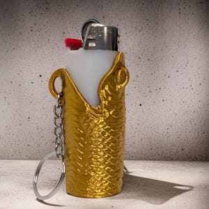 Pu&ograve; includere: Porta accendino dorato con portachiavi. Il porta accendino ha la forma di un pesce e una superficie testurizzata. L'accendino &egrave; bianco e ha un pulsante rosso.