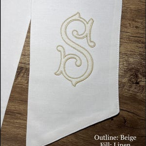 May include: White fabric with a beige embroidered letter "S". The embroidery has a decorative, ornate design. The text "Outline: Beige Fill: Linen" is visible in the lower right corner. The fabric is on a wooden surface.