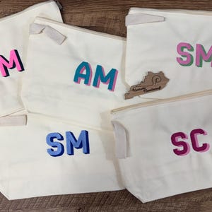 May include: Five off-white canvas zipper pouches featuring embroidered two-letter monograms in various color combinations including pink, black, teal, blue, green, and purple. A small wooden cutout shaped like Kentucky is placed among the pouches.