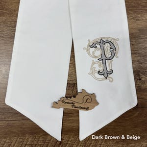 May include: A white fabric runner featuring an embroidered letter P in dark brown and beige with ornate scrollwork. A wooden cutout shaped like the state of Kentucky is placed on the fabric, displaying the text Kentucky Needlecraft.