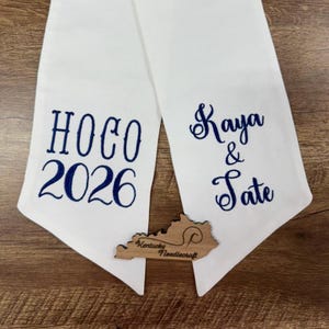 May include: A white fabric sash featuring dark blue embroidered text. The left side displays HOCO 2026, while the right side shows Kaya & Tate. A small wooden cutout shaped like the state of Kentucky with the text Kentucky Needlecraft is included.