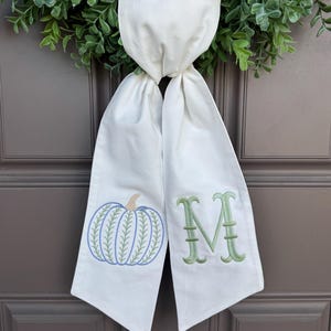 May include: A white fabric door hanger featuring an embroidered blue pumpkin with a tan stem on the left side and a large green embroidered letter M on the right side. The fabric is knotted at the top and hangs down in two separate panels.