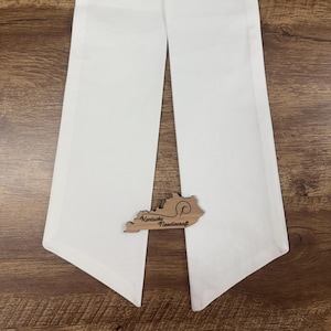 May include: Two white fabric sashes with pointed ends, arranged vertically. A wooden cutout in the shape of Kentucky with the text "Kentucky Needlecraft" is placed between the sashes. The background is a wooden surface.