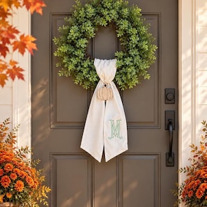 May include: A round green leaf wreath hangs on a dark brown door. A white fabric sash is draped over the center of the wreath, featuring a small wooden pumpkin cutout at the top and a large green embroidered letter M on the lower section.