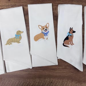 May include: White decorative towels embroidered with various dog breeds, including a black Labrador, a Dachshund, a Corgi, a German Shepherd, and a Golden Retriever. Each dog has a colorful bow.