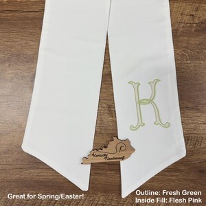 May include: White graduation stole with a large embroidered letter "K" in fresh green outline and flesh pink fill. A wooden Kentucky Needlecraft logo is at the bottom. The stole is displayed on a wooden surface.