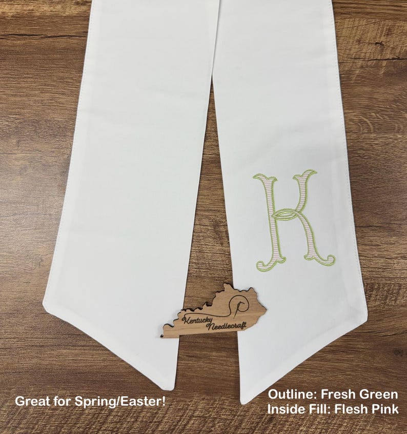 May include: White graduation stole with a large embroidered letter "K" in fresh green outline and flesh pink fill. A wooden Kentucky Needlecraft logo is at the bottom. The stole is displayed on a wooden surface.