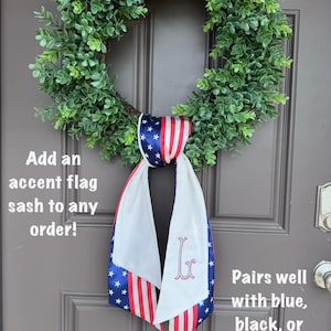 May include: A green wreath hangs on a brown door, adorned with a patriotic flag sash. The sash features a blue star pattern, red and white stripes, and a monogram. Text on the image reads: "Add an accent flag sash to any order!" and "Pairs well with blue, black, or grey color letters too!"