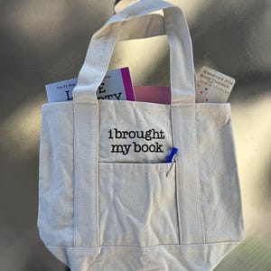 May include: A beige canvas tote bag with two handles and an external front pocket. The text "i brought my book" is embroidered in black on the front. Inside the bag are books and a card labeled "JENNIFER'S 2026 BOOK TRACKER". A blue pen is tucked into the pocket.