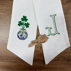 May include: White fabric towel with green shamrocks in a blue and white vase. A wooden Kentucky-shaped cutout with "Kentucky Needlecraft" is placed on the towel. A green letter "J" is embroidered on the right side.