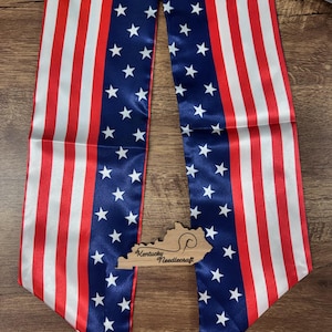 May include: A patriotic sash featuring the American flag design with red and white stripes and a blue field of white stars. A wooden cutout of Kentucky with "Kentucky Needlecraft" is placed on the sash.