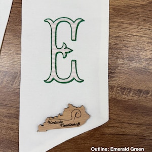 May include: White fabric banner with an embroidered letter "E" in emerald green outline and flesh pink fill. A wooden Kentucky-shaped embellishment with "Kentucky Needlecraft" is attached. The banner is displayed on a wooden surface.