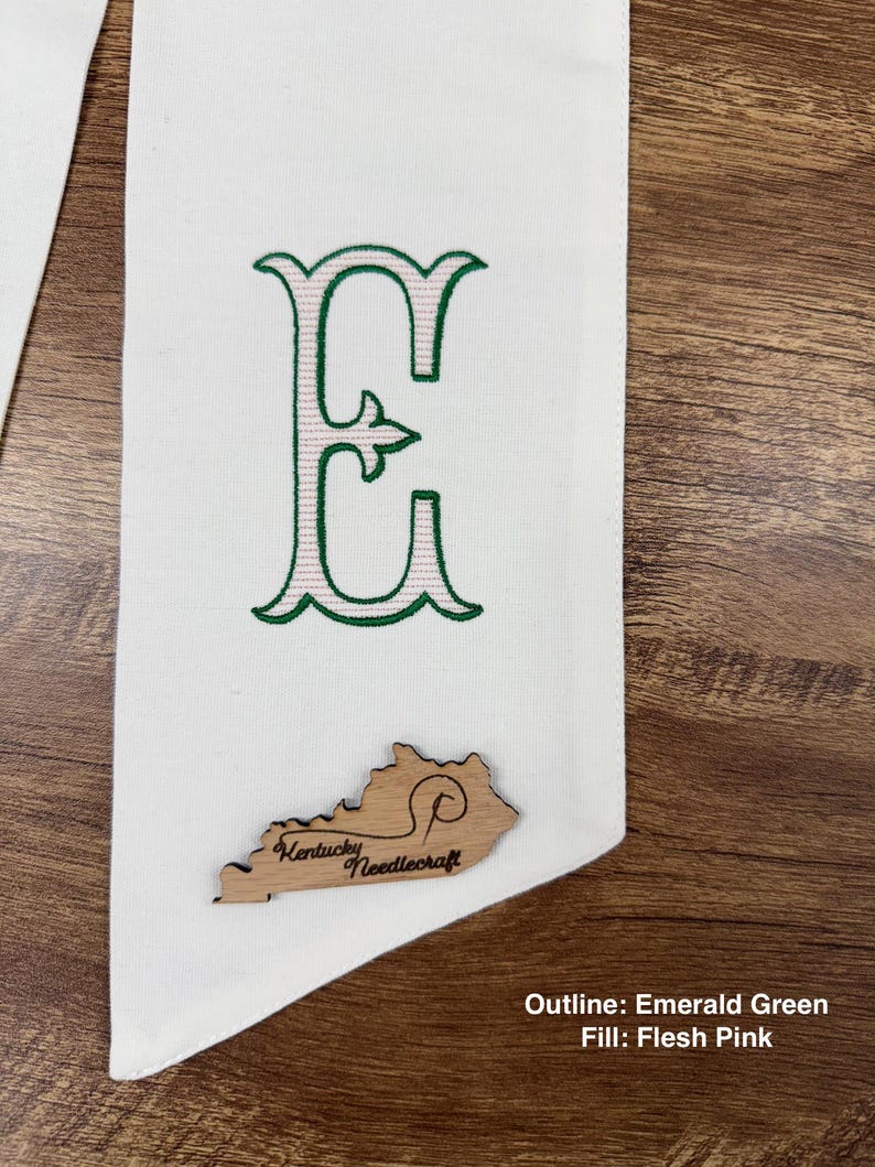 May include: White fabric banner with an embroidered letter "E" in emerald green outline and flesh pink fill. A wooden Kentucky-shaped embellishment with "Kentucky Needlecraft" is attached. The banner is displayed on a wooden surface.
