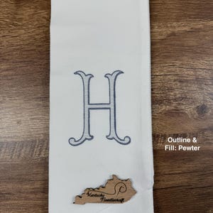 May include: White hand towel with a large embroidered letter "H" in blue. A wooden Kentucky-shaped embellishment with the text "Kentucky Needlecraft" is attached. The towel is folded and displayed on a wooden surface.