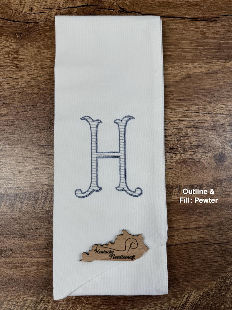 May include: White hand towel with a large embroidered letter "H" in blue. A wooden Kentucky-shaped embellishment with the text "Kentucky Needlecraft" is attached. The towel is folded and displayed on a wooden surface.