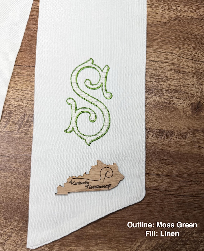 May include: White fabric sash with a green embroidered letter "S" and a wooden Kentucky-shaped emblem. The emblem has the text "Kentucky Needlecraft". The outline of the letter is moss green, and the fill is linen.