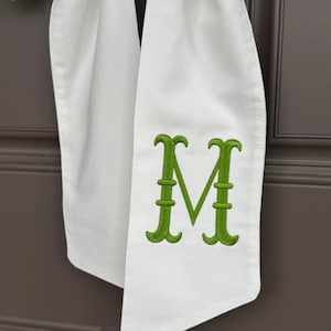 May include: White fabric door hanger with a green embroidered letter "M". The fabric is folded and hanging against a brown door. The letter is in a decorative font.
